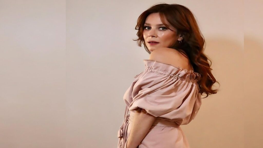 Anna Friel in a blush off-the-shoulder dress with puff sleeves gazes over her shoulder against a warm beige backdrop.