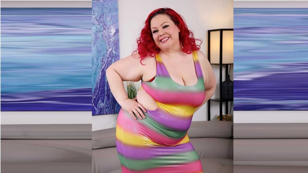 April Flores with bright red curls wearing a colorful bodycon dress, smiling and posing in a living room with blue art.