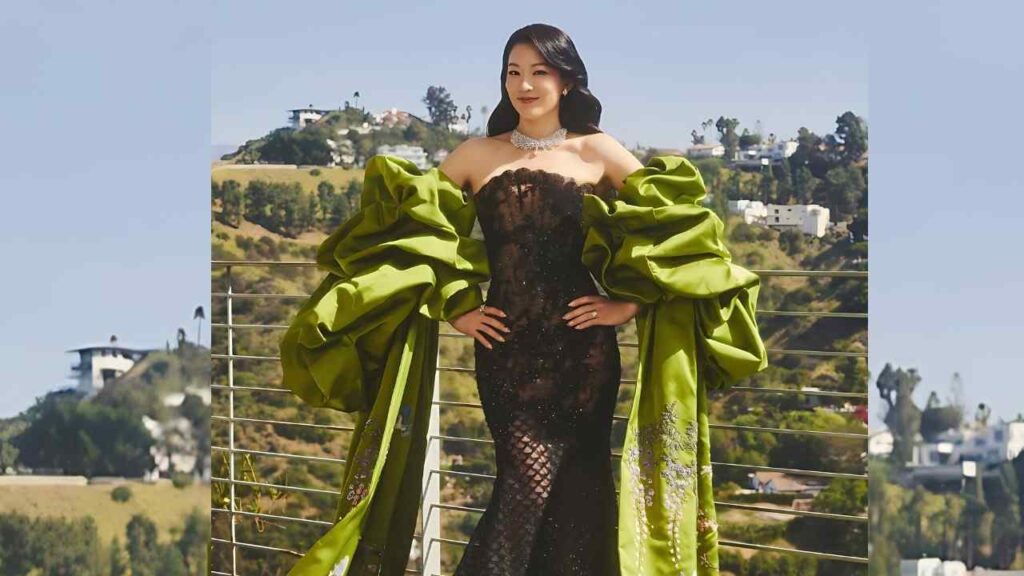 Arden Cho in a black lace off-shoulder gown with oversized lime-green sleeves, posing on a balcony above hillside homes.