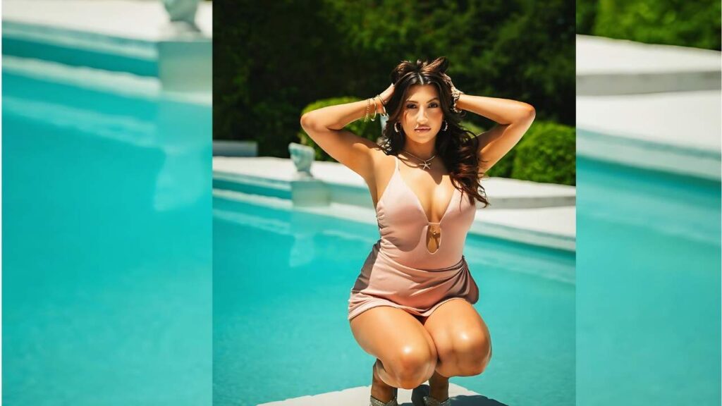 Ashley Blue kneels by a turquoise pool in a blush pink swimsuit, hands in her hair, with white steps and green hedges behind.