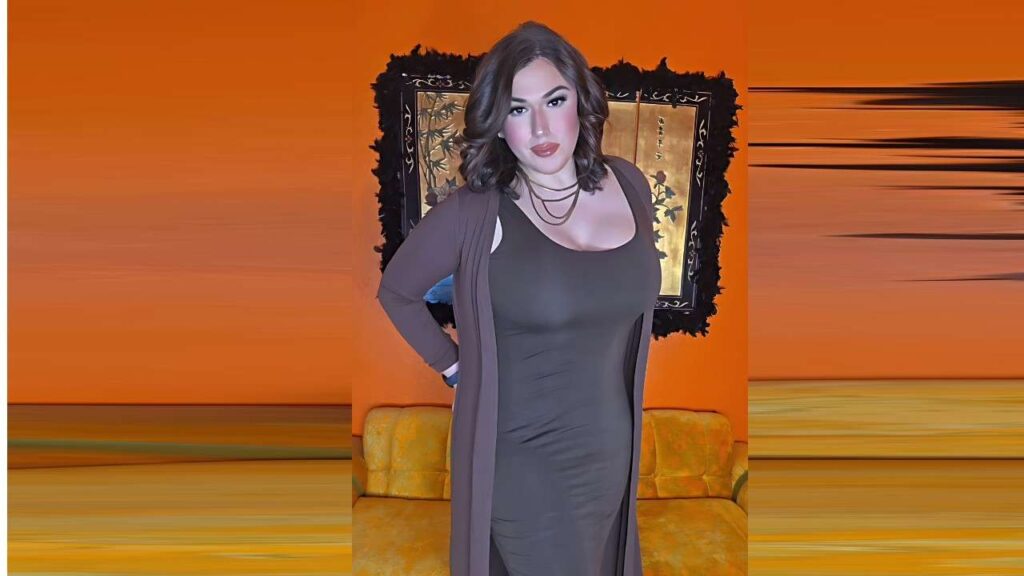 Aspen Brooks with long dark hair in a fitted gray dress and brown cardigan, hands on hips, before an orange wall with a mirror.
