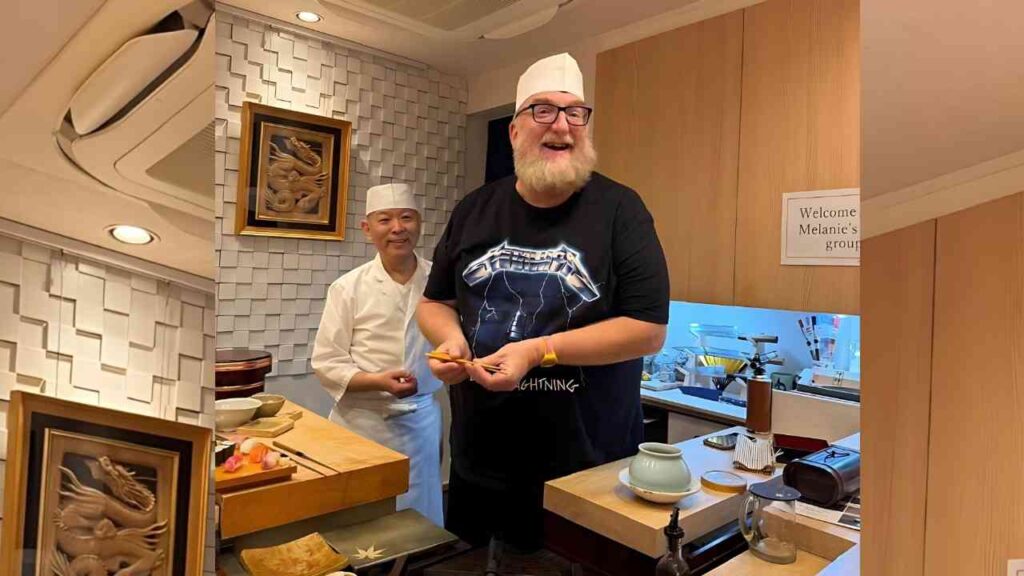 Brian Posehn in a white chef hat and black Metallica tee smiles, holding chopsticks beside a cook in a white uniform in a modern kitchen.