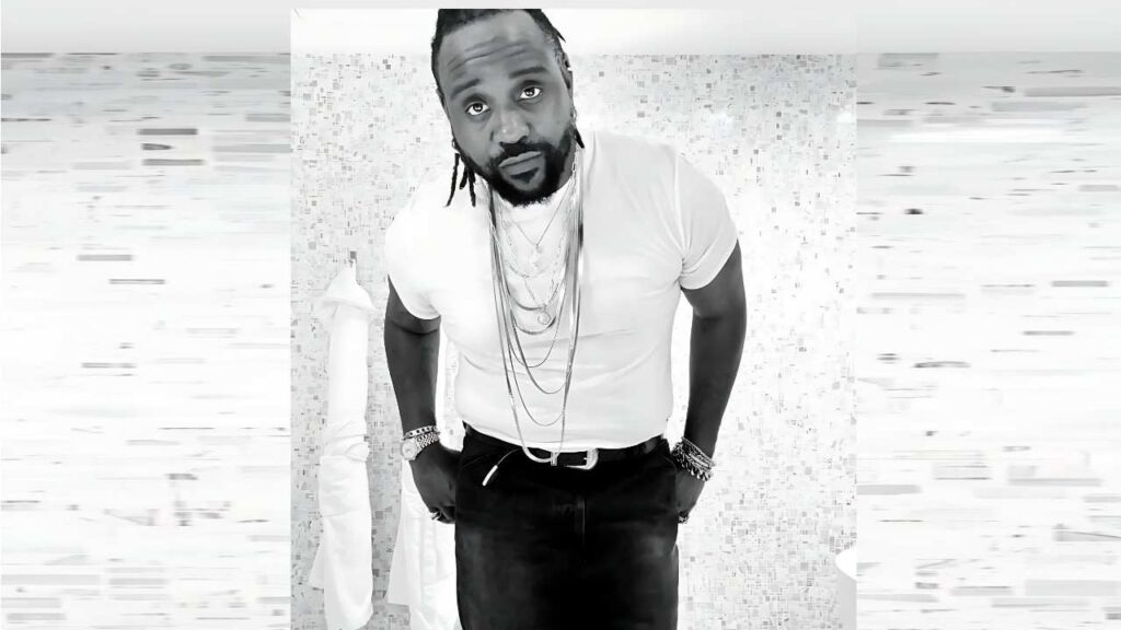 Brian Tyree Henry with dreadlocks in a white tee, wearing chains, hands in pockets, before a tiled wall.