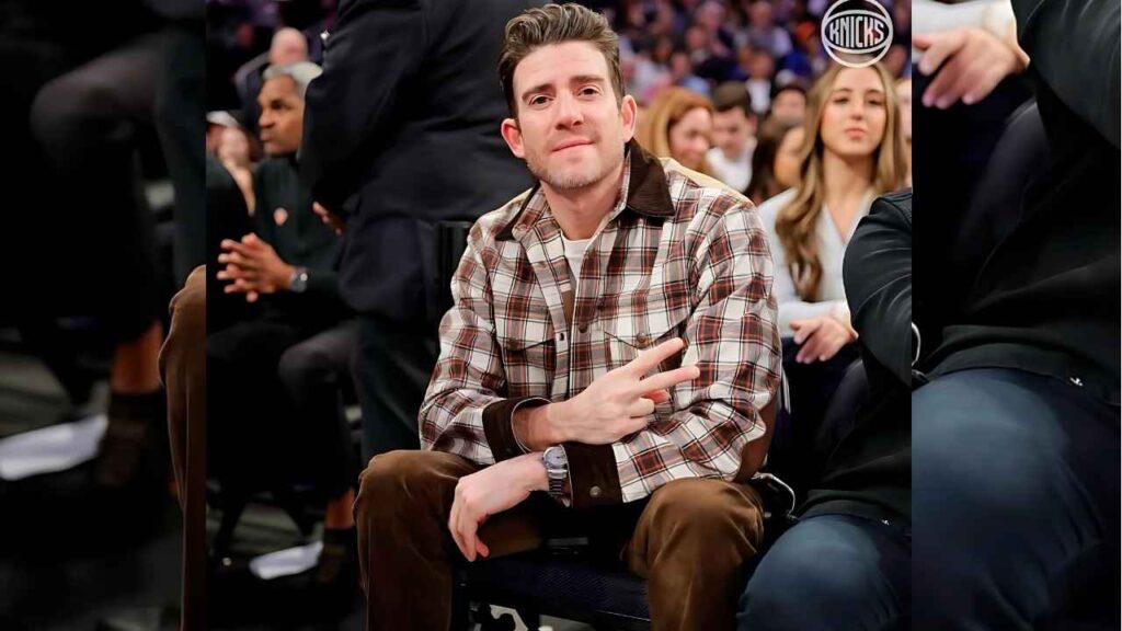 Bryan Greenberg in a brown plaid shirt and brown pants, flashing a peace sign, seated at a basketball game with a Knicks logo in the top-right corner.