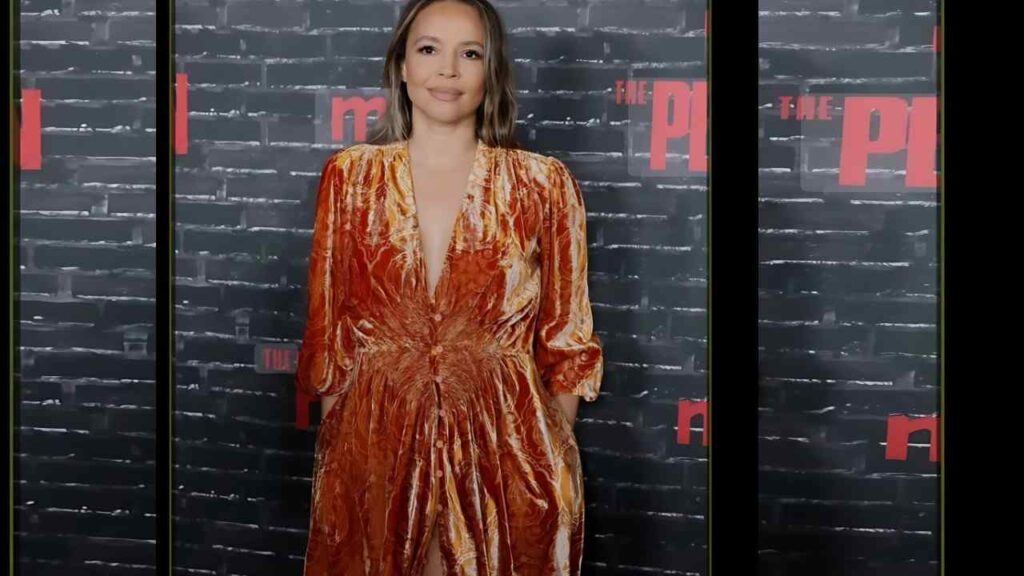 Carmen Ejogo in orange velvet dress with a deep V-neck and gathered waist stands before a dark brick backdrop with red THE PL.