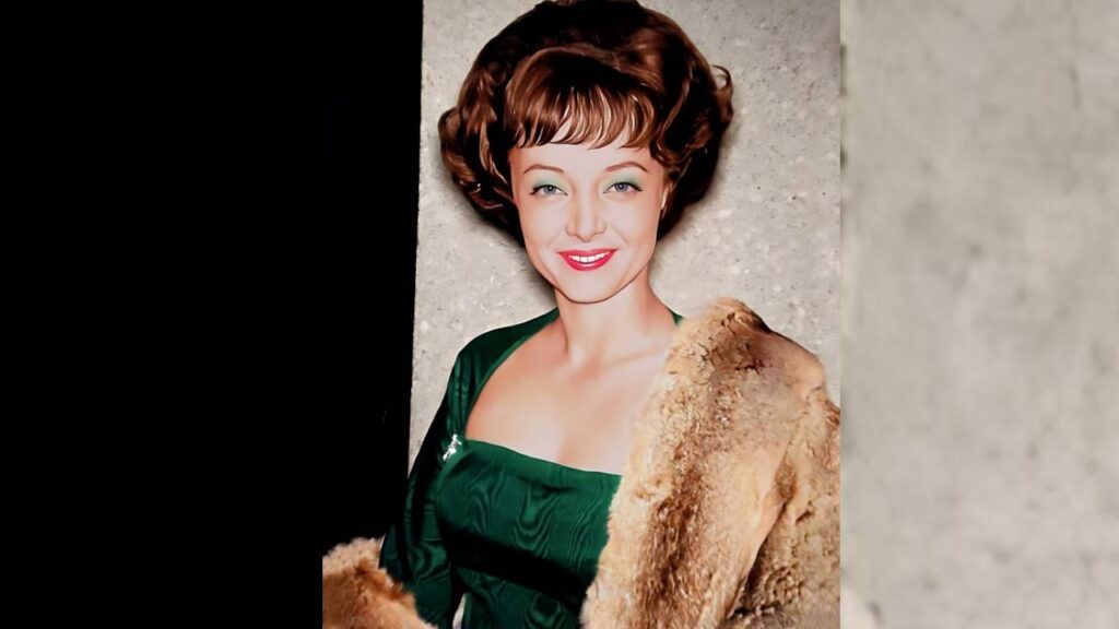 Carolyn Jones with short brown curls wearing a green satin dress and a fur coat draped over one shoulder against a grey wall.