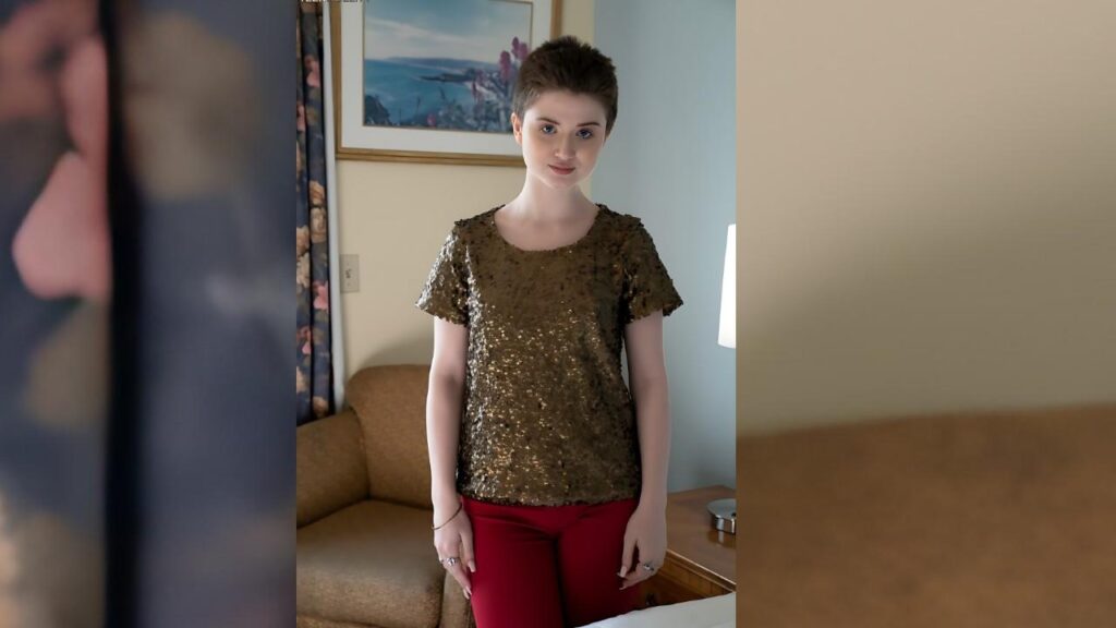 Celestia Vega with short hair stands in a living room, wearing a gold sequin top and red pants, smiling softly.