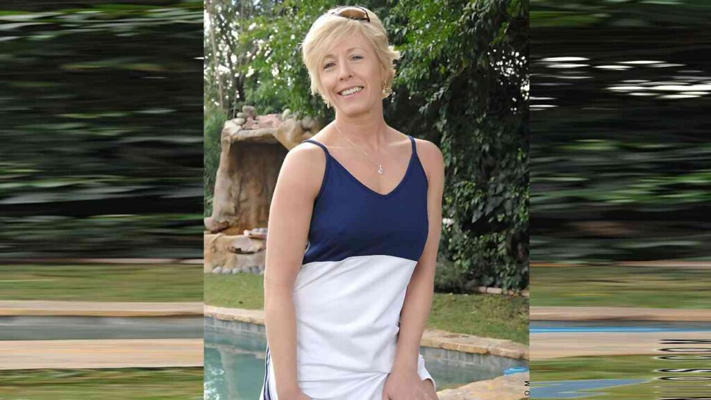 Chanel Carrera in navy and white sleeveless top, sunglasses on her head, stands by a pool with leafy greenery behind.