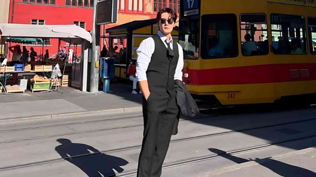 Choi Jin-hyuk in a black waistcoat and sunglasses stands on a sunny city street with a coat slung over his arm, beside a yellow tram.