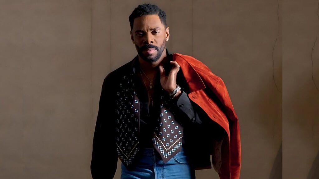 Colman Domingo with a studded black vest over a dark shirt, blue jeans, and a red jacket draped over his shoulder, posing against a beige backdrop.