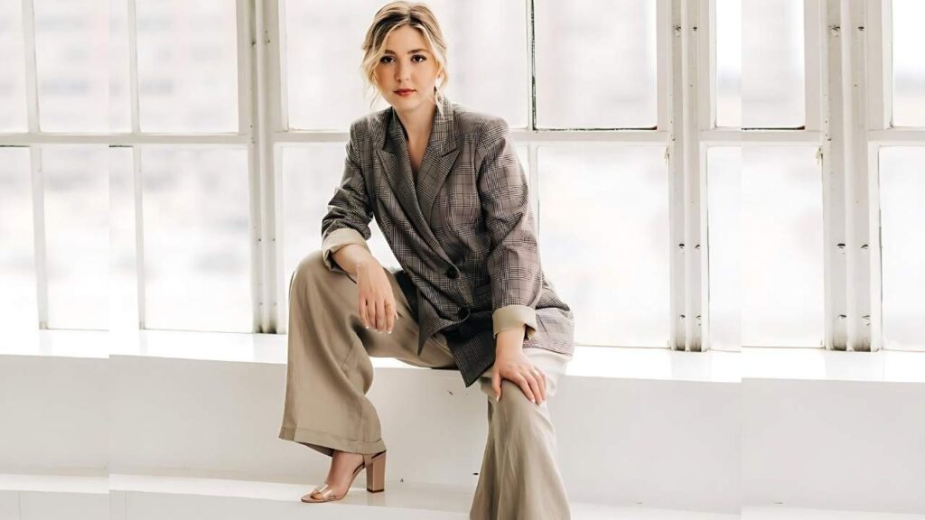 Cozi Zuehlsdorff sits on a white ledge by large windows, wearing a plaid blazer and beige wide-leg trousers with tan sandals.
