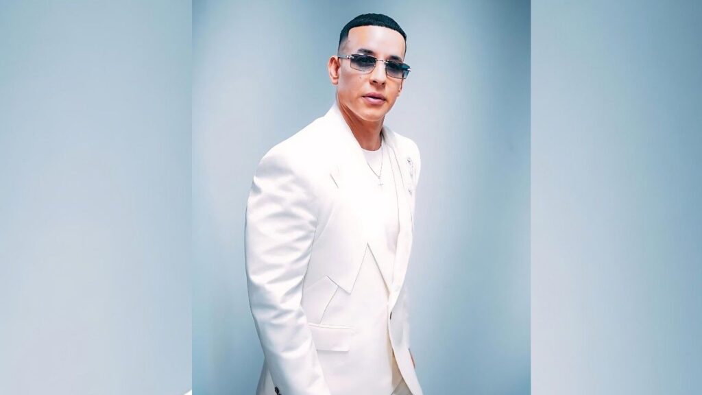Daddy Yankee in a white suit and sunglasses stands against a pale blue gradient backdrop, looking at the camera with expression.