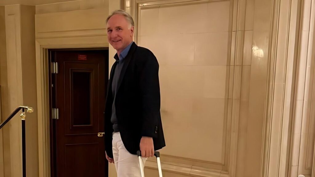 Dan Brown in a dark blazer, blue shirt and white pants stands in a marble hotel hallway, holding a suitcase handle.