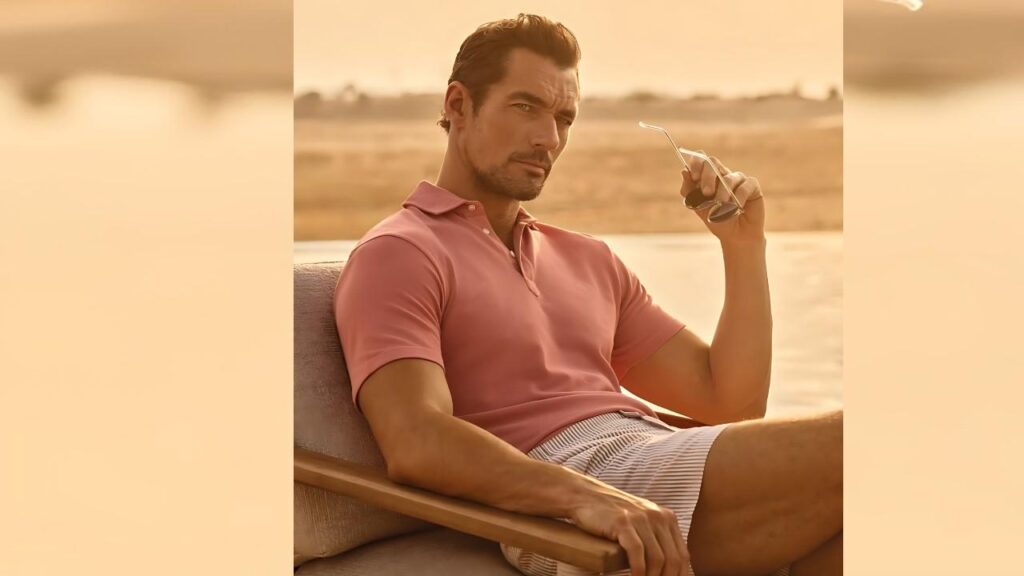 David Gandy in a pink polo and striped shorts lounges on a cushioned outdoor sofa by a sunset-lit lake, holding sunglasses.