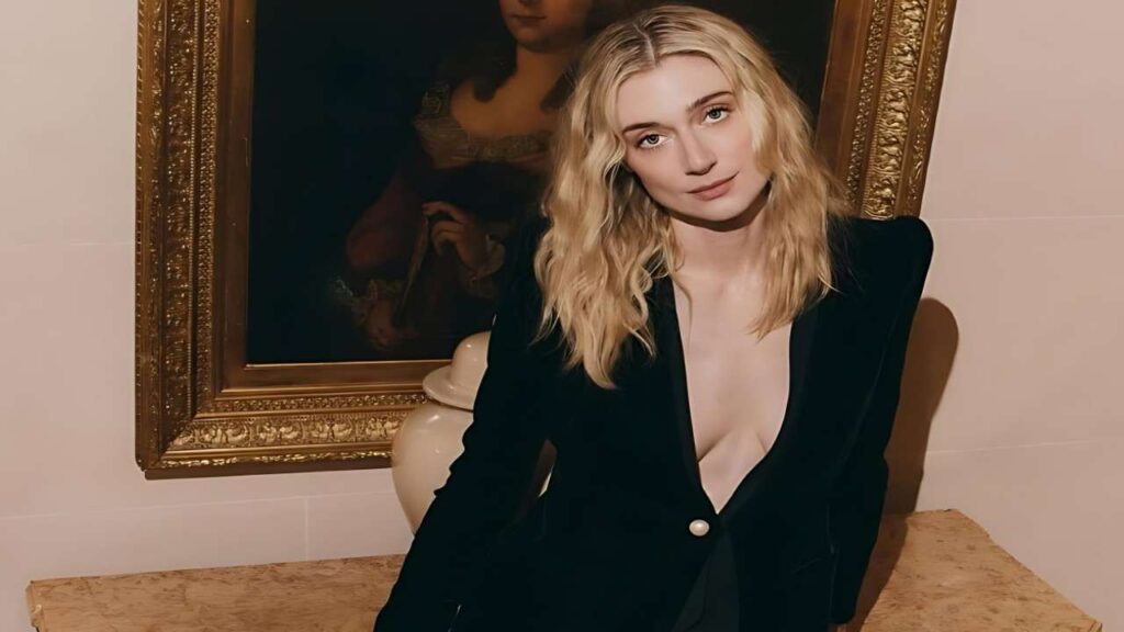 Elizabeth Debicki with wavy hair in a plunging black blazer sits at a marble counter, vase and gold frame painting behind her.