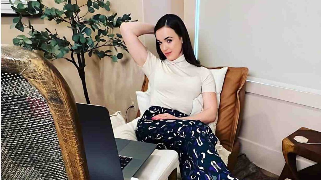 Elizabeth Henstridge with dark hair sits on a cream sofa, white top and navy pants, laptop on table, wicker chair and plant nearby.