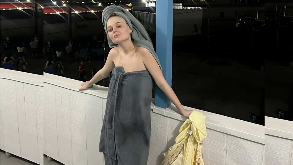 Emily Alyn Lind in a gray towel wrap and turban head wrap leans on a white tiled railing beside a blue post, with a yellow towel draped nearby.