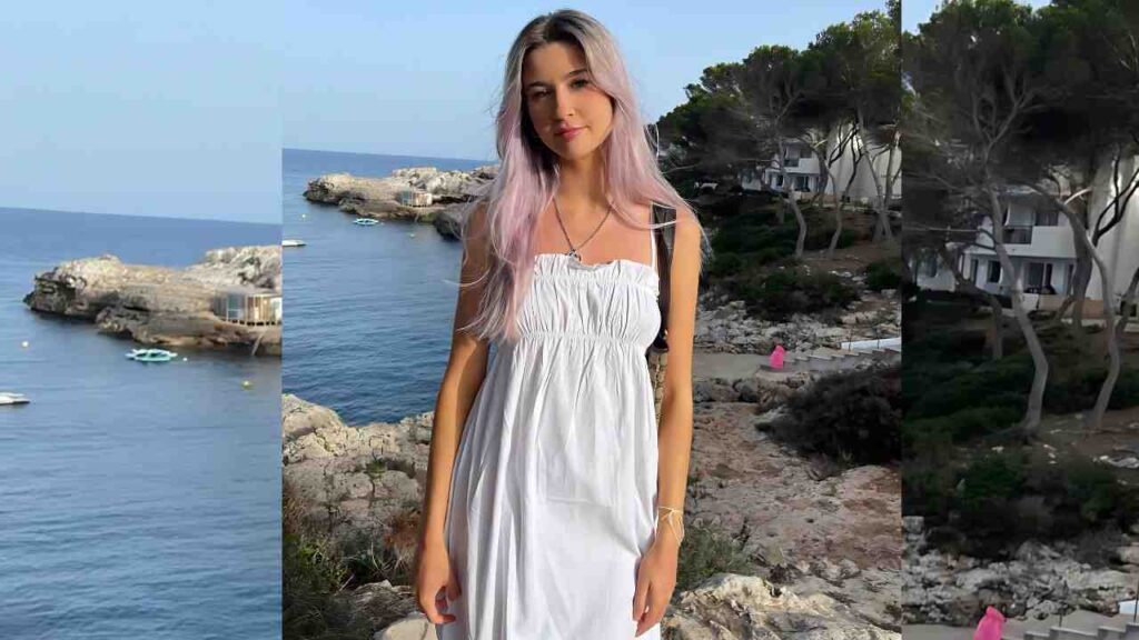 Emily Jade with pastel pink hair in a white smocked dress stands on a rocky coastal overlook, blue sea and boats in view.