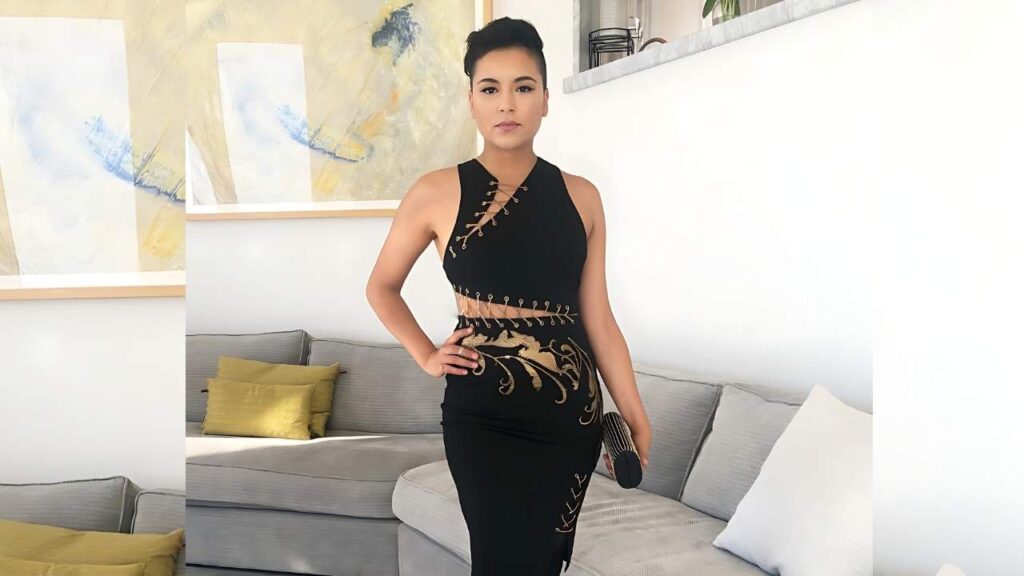 Emily Rios in a black sleeveless dress with gold accents, posing with a hand on her hip in living room with a gray sofa.