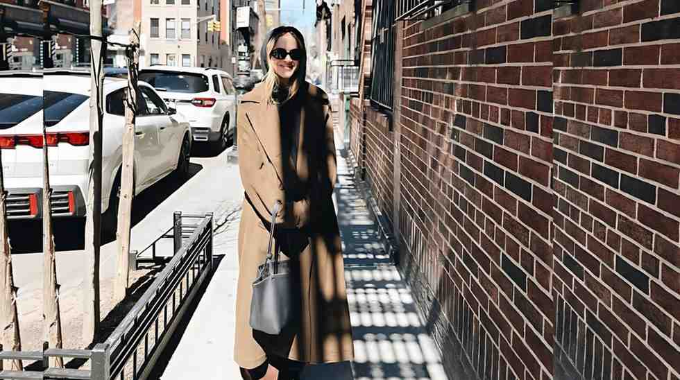 Emily Wickersham in a camel long coat, black outfit and sunglasses walks a sunlit city sidewalk, carrying a gray tote by a brick wall.
