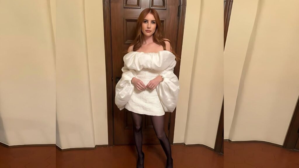Emma Roberts in a doorway wears an off-shoulder white puff-sleeve top with a short textured white skirt and black tights.