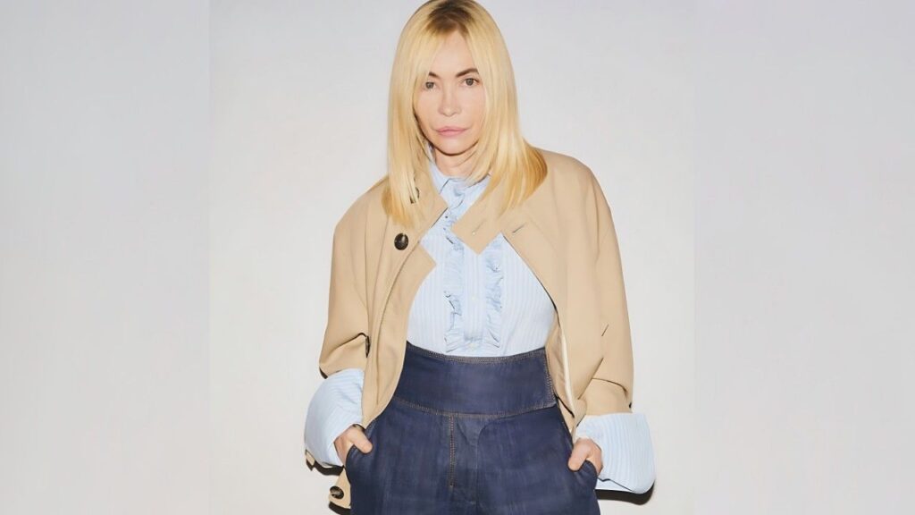 Emmanuelle Béart stands against a pale backdrop in a light blue ruffled blouse and beige cropped jacket draped over shoulders, hands in pockets.