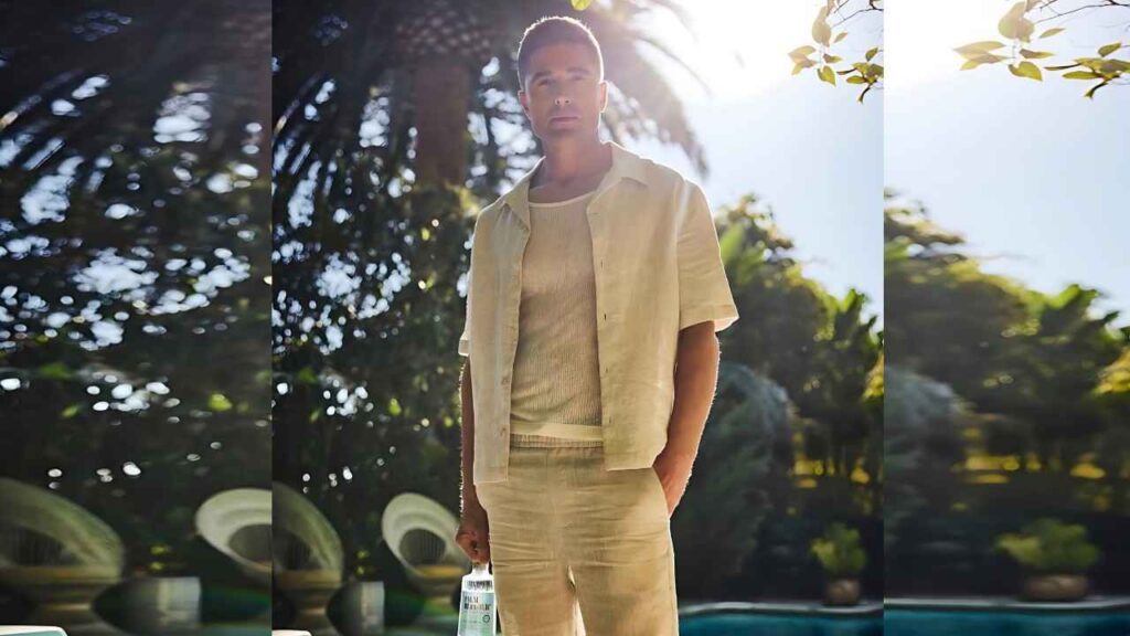 Eric Winter stands poolside in a sunny tropical yard, palm trees and greenery behind, holding a bottle.