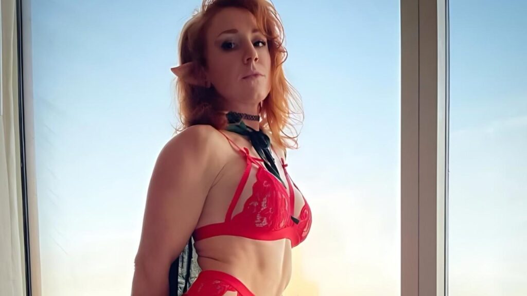 Erika Knight in red lace and a green scarf, with a black choker, poses by a sunlit window against a blue sky.