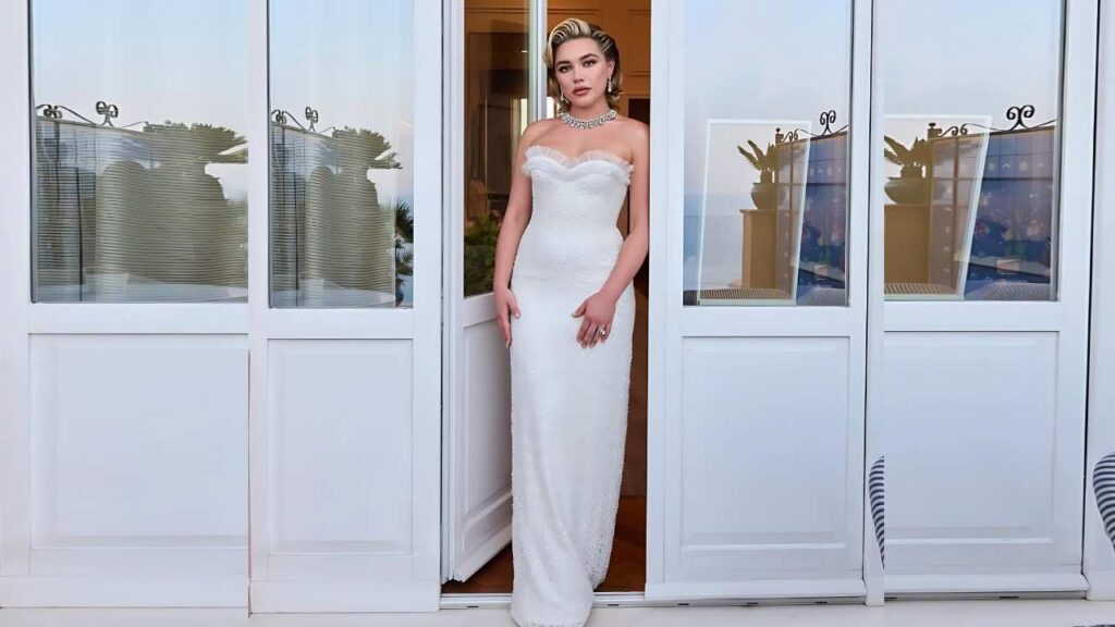Florence Pugh in a white strapless gown stands in a glass-paneled doorway, with vintage waves and sparkling necklace.