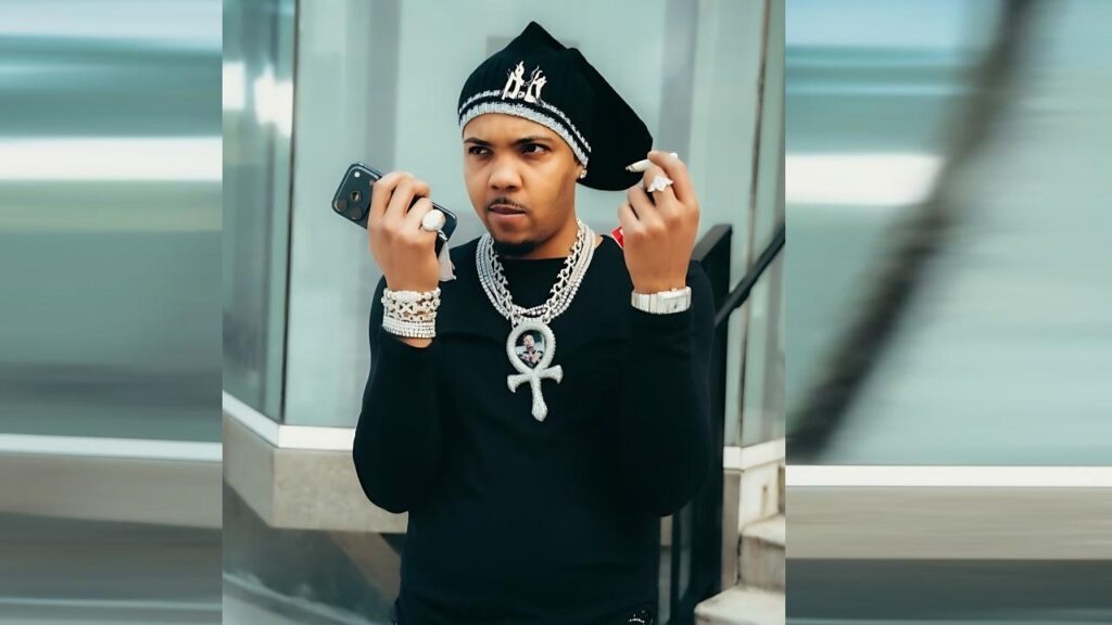 G Herbo in black shirt with a headwrap, wearing heavy diamond jewelry and an Ankh pendant, holds a phone beside a glass building.