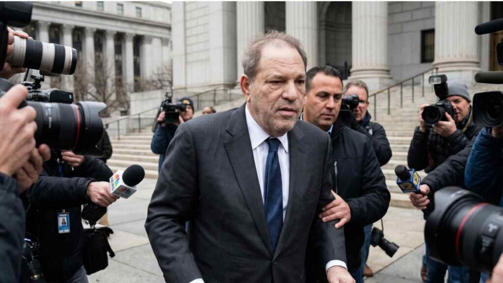 Harvey Weinstein in a dark suit and blue tie walks out as reporters and photographers swarm around him outside a grand, columned building.