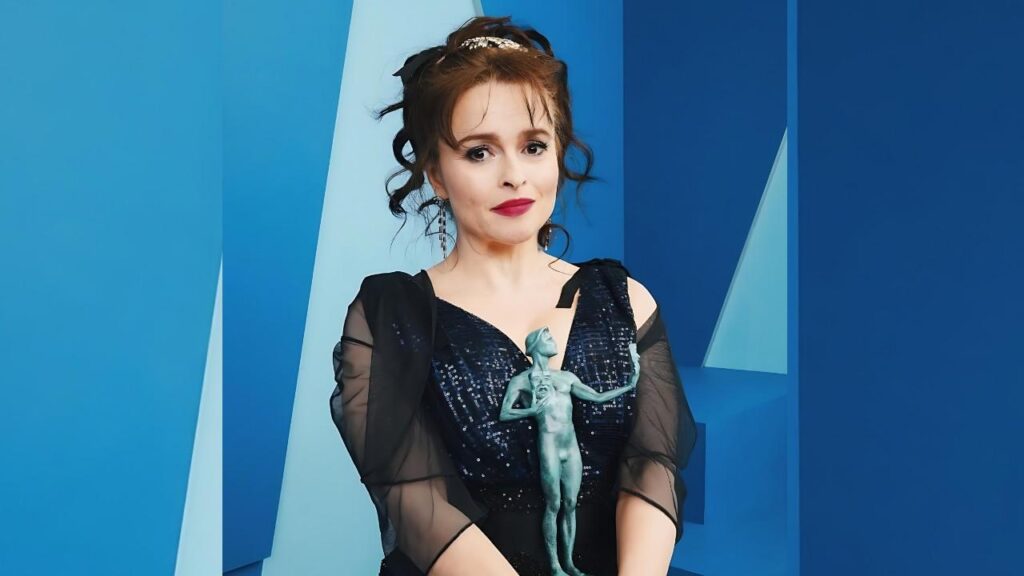 Helena Bonham in a sequined gown with sheer sleeves and red lips, wearing a teal dancer brooch, against blue backdrop.