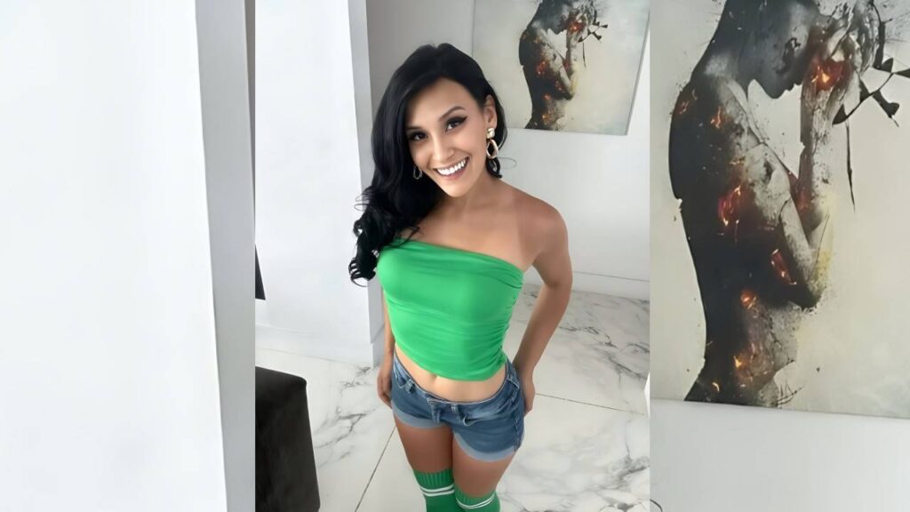 Jade Greene with black wavy hair in a bright green strapless top and denim shorts, posing in a white marble room with art.