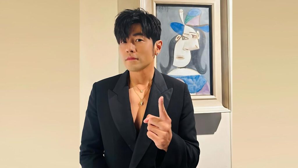 Jay Chou in a black blazer with an unbuttoned shirt and gold pendant, pointing upward beside a framed abstract painting.