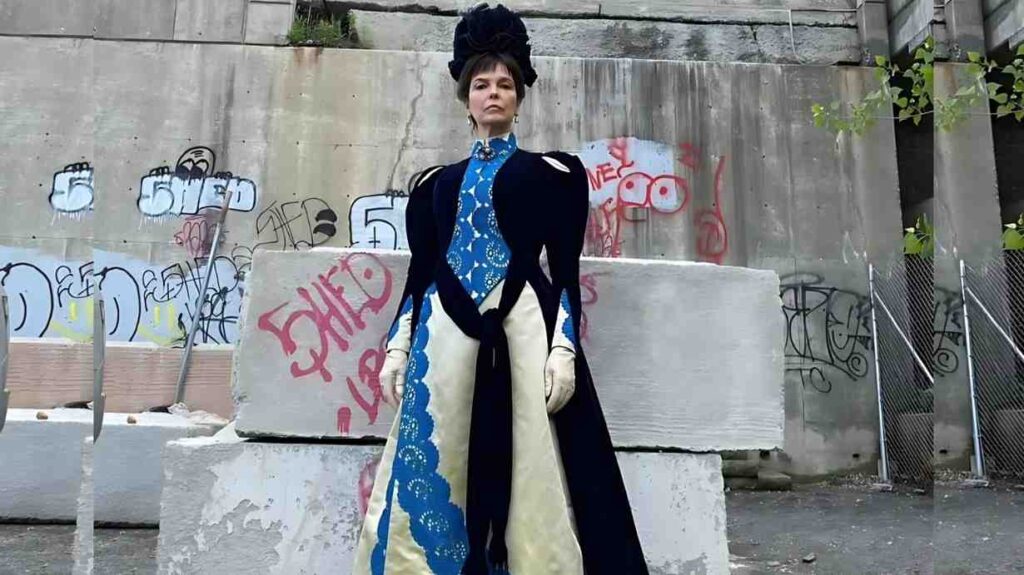 Jeanne Tripplehorn inspired gown with blue velvet cape, cream panels and lace trim, white gloves, headpiece, standing in a graffiti-covered urban yard.