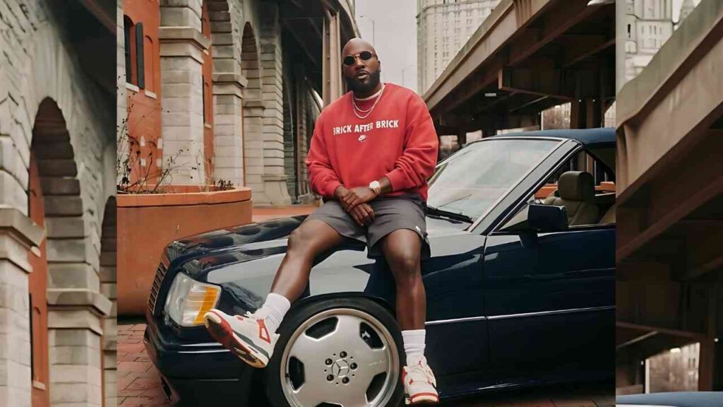Jeezy in red sweatshirt and sunglasses sits on the hood of a dark blue Mercedes, urban brick street with elevated rails.