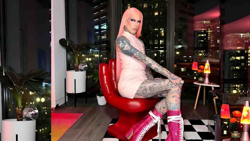 Jeffree Star in a pink dress sits sideways on a red sculptural chair, tattoos visible, in a loft with a checkered floor.