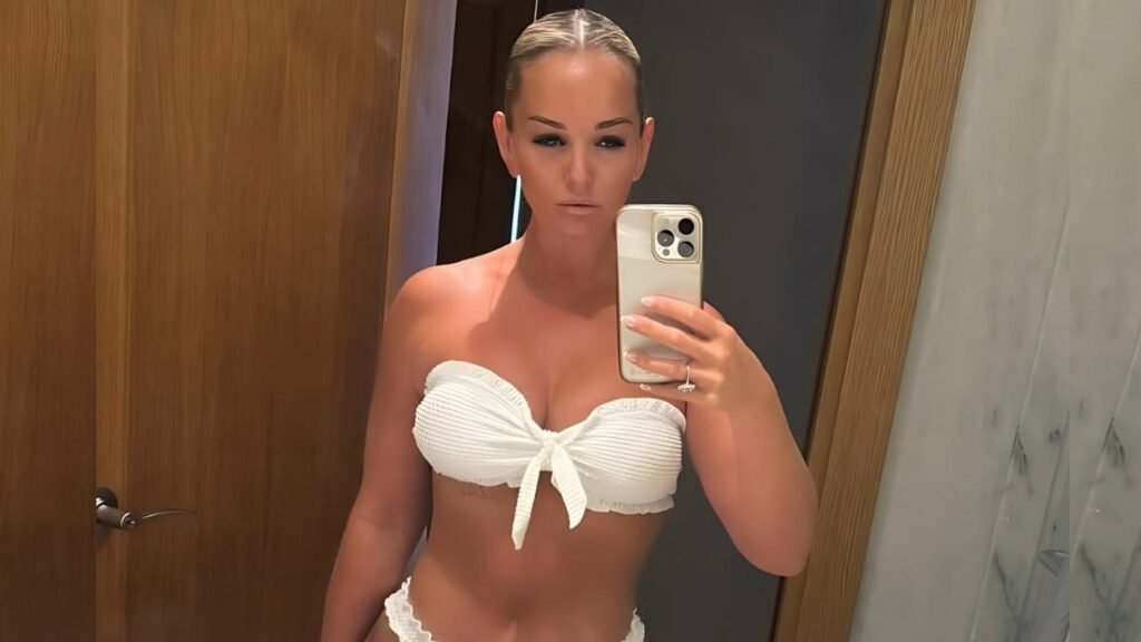 Jennifer Ellison in a white bandeau bikini with a bow, taking a mirror selfie in a wooden-and-marble bathroom.