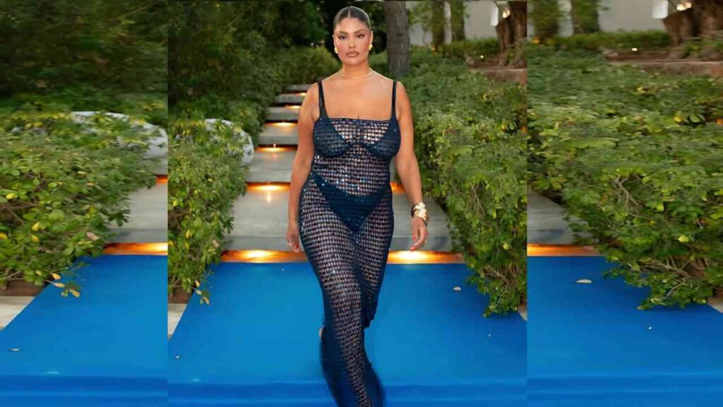 Jocelyn Corona walks a blue runway in navy crochet mesh dress, visible undergarments, gold cuff, bun, lush greenery.