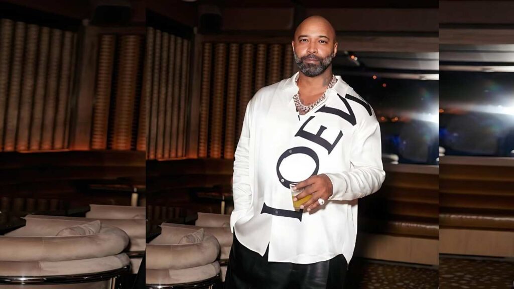 Joe Budden in a white oversized shirt with bold 'LOVE', holding a yellow drink in a dim lounge with curved seating.