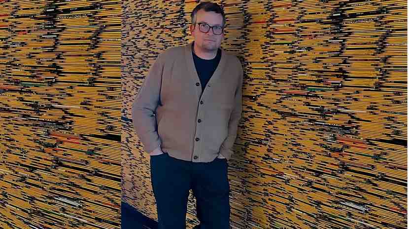 John Green wearing a tan cardigan over a black shirt and glasses stands with hands in pockets against a yellow striped backdrop.