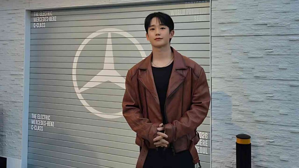 Jung Hae-in in a brown leather jacket stands with hands clasped before a gray shutter bearing a large Mercedes-Benz logo.