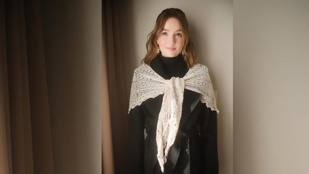 Kaitlyn Dever in a black turtleneck, wearing a cream crochet shawl tied at the front, with wavy hair.