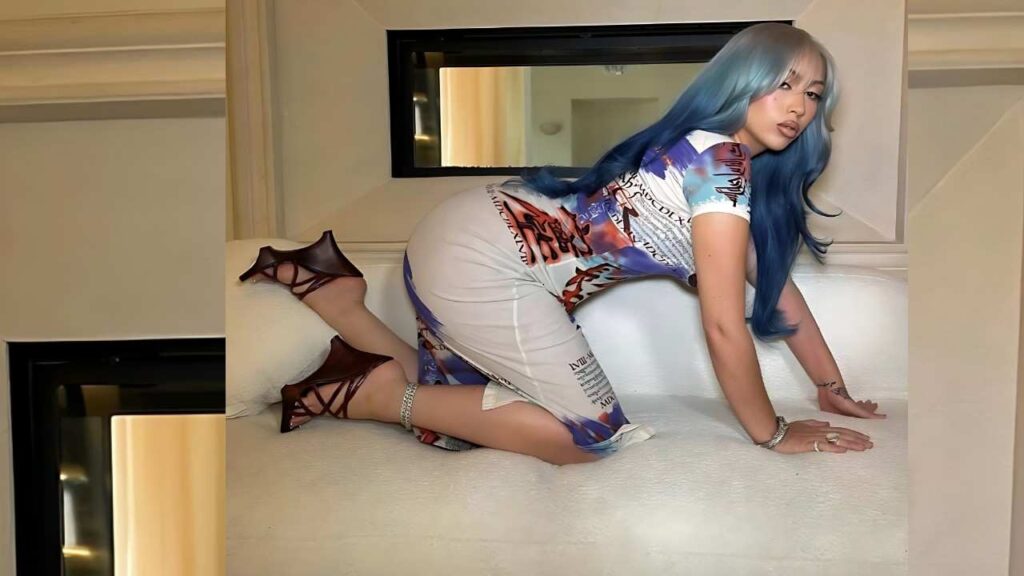 Kali Uchis in a graphic print bodycon dress kneels on a white couch, wearing strappy heels, hands on cushions.