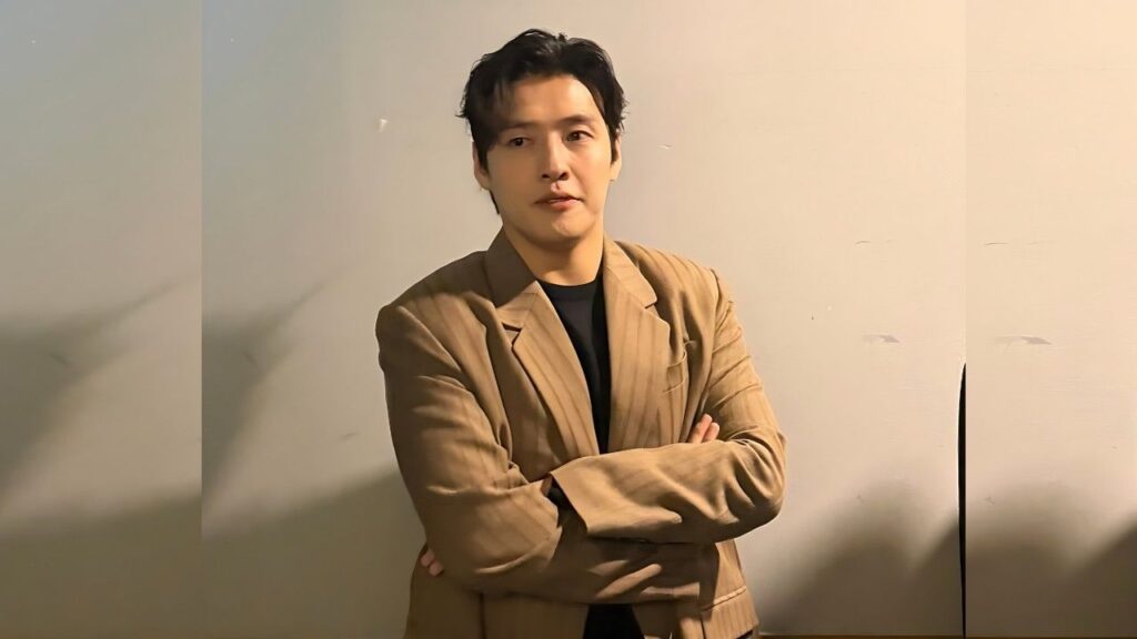 Kang Ha-neul with dark hair stands against a pale beige wall, arms crossed, wearing a tan blazer over a black shirt.