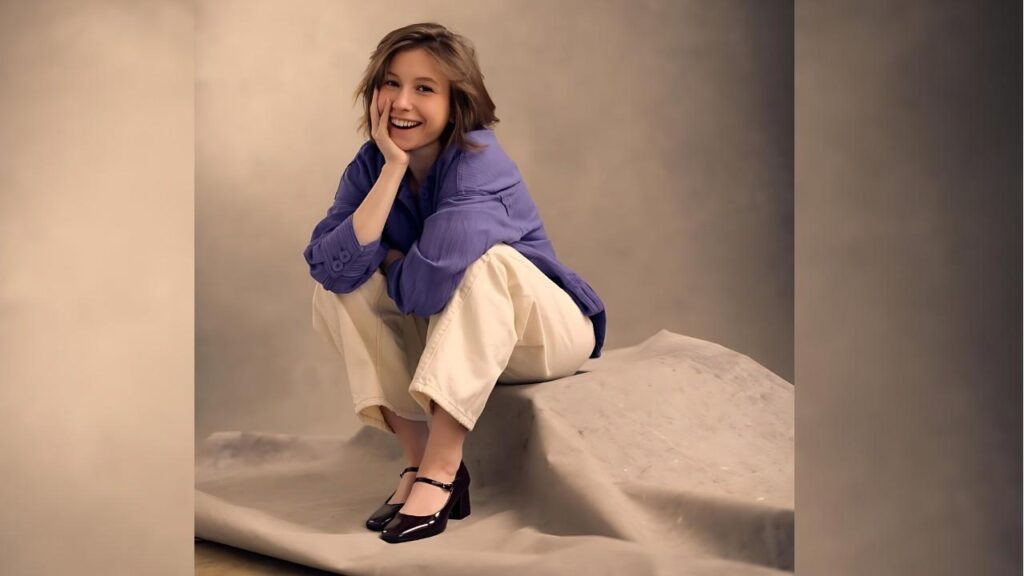 Katelyn Nacon in blue blouse and cream pants sits on a stone prop, resting her chin on her hand, against a beige backdrop