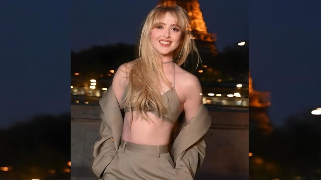 Kathryn Newton in a beige two-piece suit poses at night, smiling, jacket draped off shoulders, with city lights and a lit tower behind.