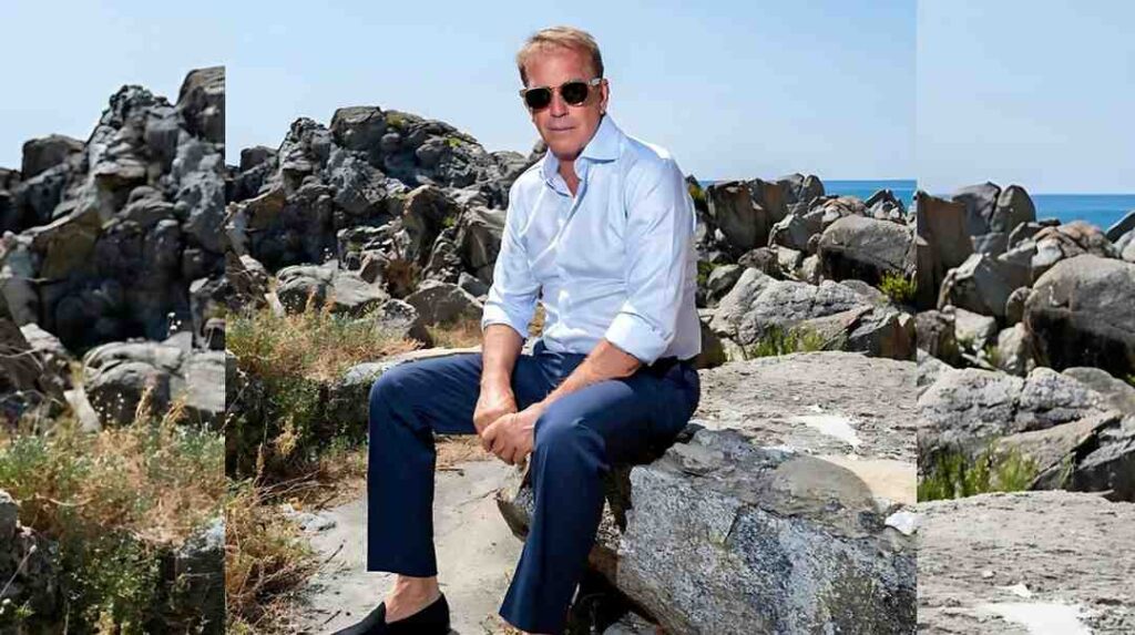 Kevin Costner in a light blue shirt and navy pants sits on a rock by the rocky shoreline, with sunglasses and sea in the background.