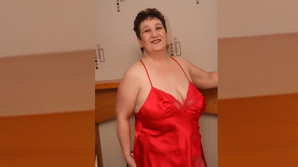 Kinky Carol with short dark hair in red satin with lace, standing by a wooden counter and leaning on it.