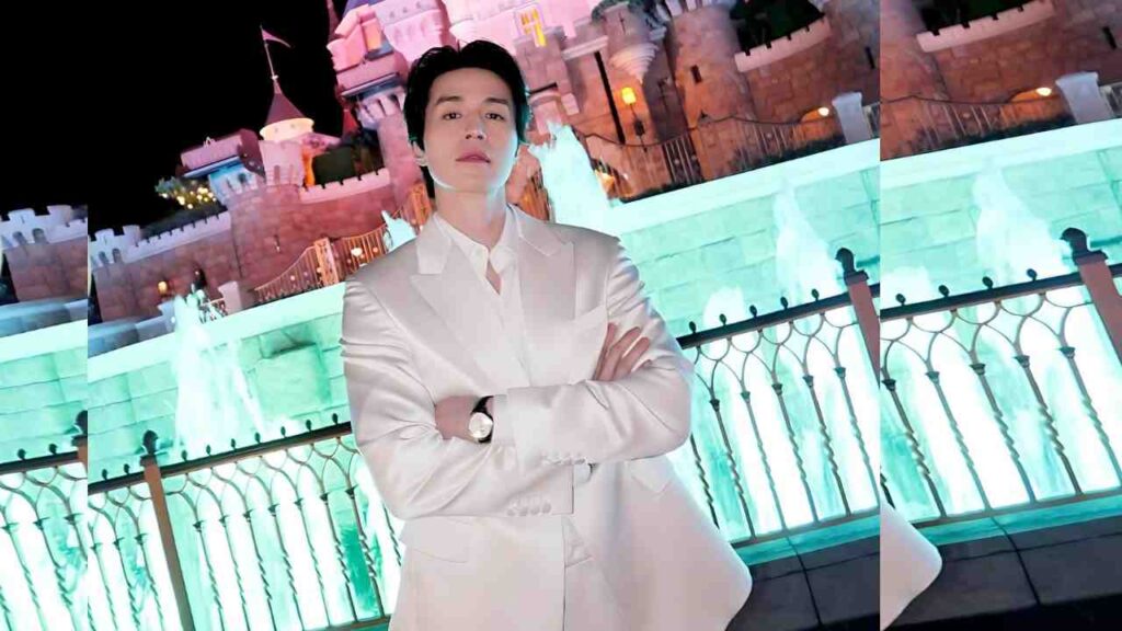 Lee Dong-wook in a white suit with arms crossed stands before a colorful illuminated castle and turquoise-lit fountain railing.